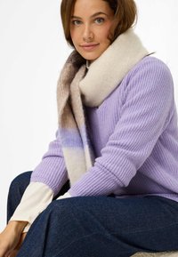 Lila gestrickter Pullover mit gerippter Textur. Das Model trägt einen dicken creme- und beigen gestreiften Schal. Dunkelblaue Denim-Hosen. Neutraler Hintergrund.