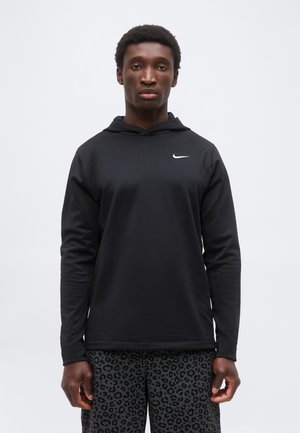 Joven con sudadera con capucha negra de Nike y pantalones grises oscuros con estampado de leopardo, de pie frente a un fondo claro y simple.