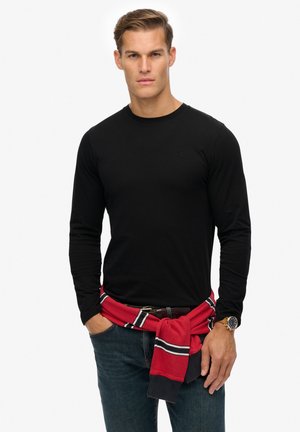 Camiseta negra de manga larga con cuello redondo, que presenta un pequeño emblema. Combinada con jeans oscuros y un sudadera a rayas rojas atada a la cintura.