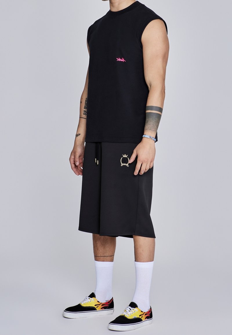 Homme portant un débardeur noir, un short long noir avec un blason, des chaussettes blanches et des baskets noires avec des flammes jaunes et rouges.