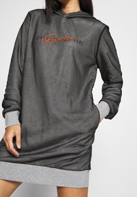 Vestido sudadera con capucha en tejido gris oscuro con puños en gris más claro. Presenta un logo bordado en naranja y un corte relajado con una superficie ligeramente texturizada.