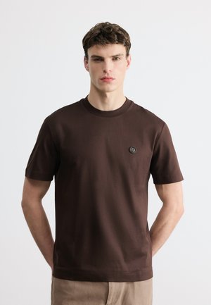 Νεαρός άντρας με κοντά καστανά μαλλιά που φοράει απλό σκούρο καφέ t-shirt με μικρό στρογγυλό λογότυπο στο αριστερό στήθος, μπεζ παντελόνια και ουδέτερο φόντο.