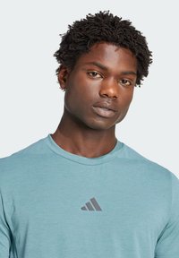Un t-shirt de sport bleu clair en tissu doux, avec un col rond et un logo Adidas gris sur la poitrine, doté de manches courtes.
