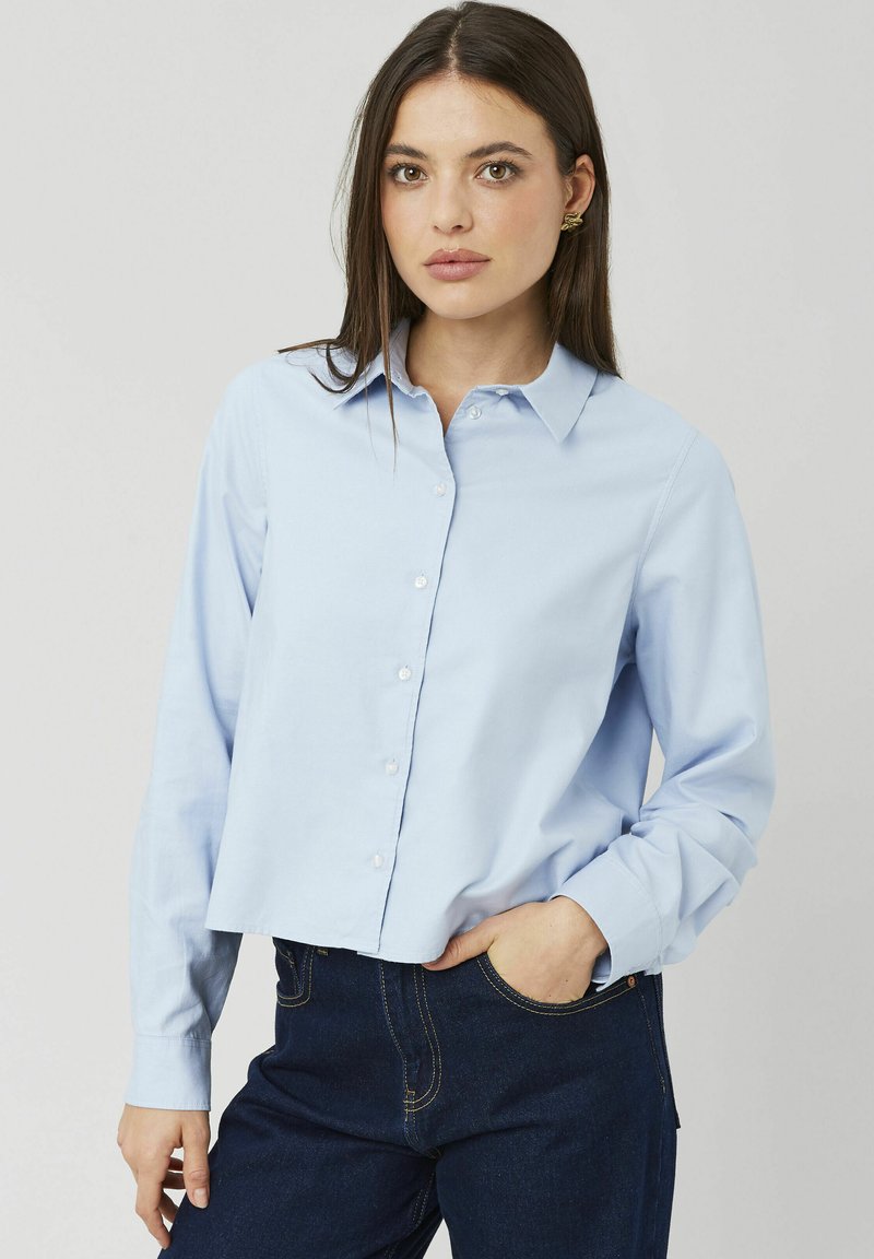Chemise boutonnée bleu clair avec col à pointes écartées, manches longues et design court, associée à un pantalon en denim bleu foncé. Texture lisse.