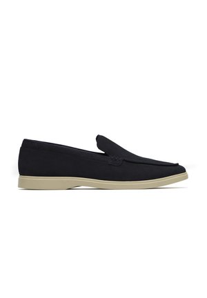 Chaussure slip-on en daim noir avec un bout arrondi, une semelle en caoutchouc beige et un design minimaliste. Dotée d'un seul point de couture accentué.