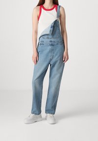 Denim overalls in light blue with a single shoulder strap. The design features a pocket on the bib and wide, relaxed legs. Paired with white sneakers.