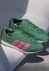 Zelené športové topánky s ružovými akcentmi, sieťovaným zvrškom, textúrovaným povrchom a bielou podrážkou; obsahujú tri ružové pruhy na boku.