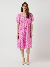 Rosa klänning täckt av rosformad tyg, med korta puffärmar, en V-ringning och ett knytbälte i midjan, i kombination med beige pumps.