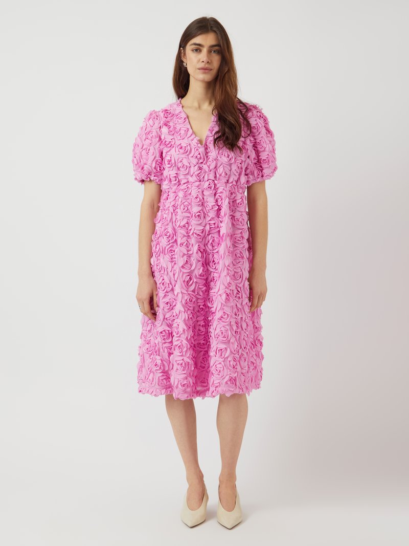 Rosa klänning täckt av rosformad tyg, med korta puffärmar, en V-ringning och ett knytbälte i midjan, i kombination med beige pumps.