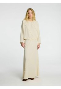 Beige to-delt sæt med en langærmet cropped top med en syet diagonalt mønster og en matchende maxi-nederdel, begge i et blødt materiale.