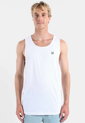 Homme blond aux yeux bleus portant un débardeur blanc uni sans manches et un short bleu clair, debout devant un fond uni.