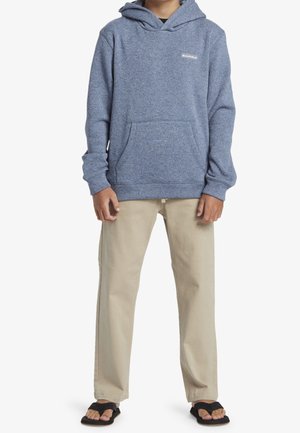 Personne portant un sweat à capuche bleu, un pantalon beige et des tongs noires, debout devant un fond blanc.