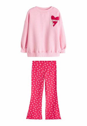 Rosa Sweatshirt mit langen Ärmeln, rotem Herz und dem Schriftzug „L'AMOUR EST BON“, kombiniert mit roten Schlaghosen, die mit kleinen weißen Herzchen gemustert sind.