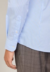 Lichtblauw en wit gestreepte lange mouwen shirt met knoopmanchetten, met een enkele zak en een rechte zoom. Grijze geruite broek zichtbaar.
