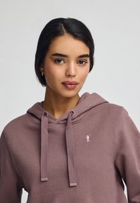 Sudadera con capucha en color malva, con cordón ajustable, fabricada en un tejido suave y un pequeño logotipo bordado en el pecho. Textura suave.