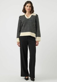 Svart och vit mönstrad stickad tröja med V-hals och krämfärgad krage, matchad med svarta vida byxor och svarta skor.