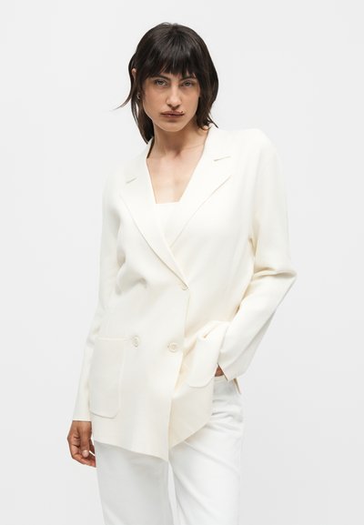 Γυναίκα με σκούρα μαλλιά που φοράει ένα κρεμ διπλόμπουκο σακάκι και παντελόνι που ταιριάζει, ποζάροντας με το ένα χέρι στην τσέπη σε απλό φόντο.