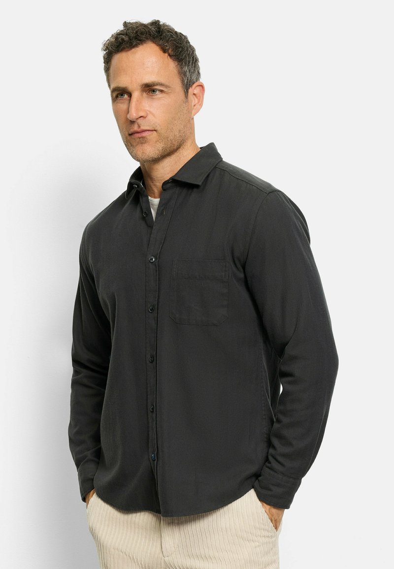 Black button-up shirt with a pocket, featuring a soft texture and long sleeves. The model wears cream-colored striped pants.