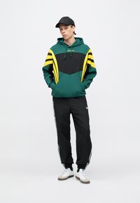 Mladý muž stojaci so založenými rukami v kapse, má na sebe zelenú, čiernu a žltú mikinu Adidas, čierne tepláky Adidas, biele tenisky a čiernu čiapku.