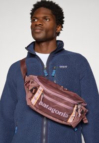 Patagonia sling bag in dark burgundy with contrasting orange and light purple accents, two zippered compartments, and an adjustable strap.