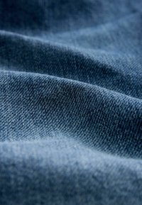 Țesătură de denim într-un albastru închis cu o suprafață texturată, prezentând o țesătură fină și variații subtile de culoare și nuanțe pe pliuri.