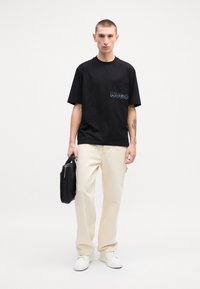 Tricou negru din bumbac cu mânecă scurtă, având logo-ul albastru "HUGO", asortat cu pantaloni crem și adidași albi, ținând o geantă neagră.