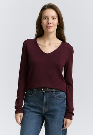 Pull en v couleur bourgogne avec des manches longues, texture côtelée, associé à un jean bleu taille haute et une ceinture noire. Design simple, coupe décontractée.