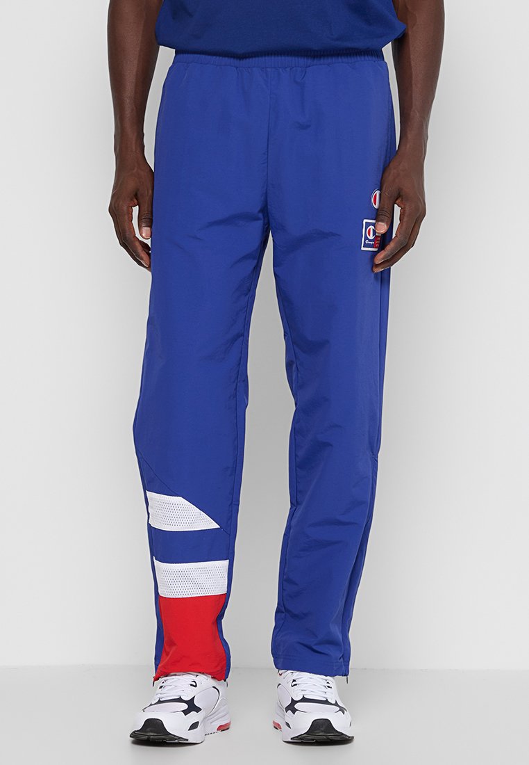 Un homme portant un pantalon de jogging bleu avec des accents blancs et rouges sur la jambe inférieure gauche, un logo Champion sur la cuisse droite, et des baskets blanches.