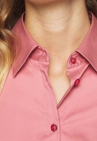 Blusa rosa con colletto, caratterizzata da una superficie liscia, con bottoni rossi e dettagli di cucitura visibili lungo la patta e il colletto.