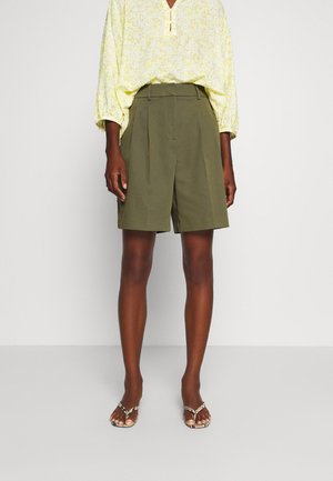 Dark-skinned person wearing loose olive-green shorts, light yellow patterned long-sleeve blouse, and open-toe sandals, standing against white background.