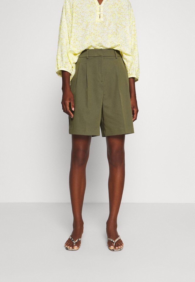 Personne à la peau foncée portant un short ample vert olive, un chemisier à manches longues jaune clair à motifs et des sandales ouvertes, debout devant un fond blanc.