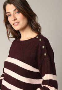 Manteau rayé bordeaux et rose clair avec une coupe décontractée, un col rond et des boutons décoratifs sur l'épaule pour un détail supplémentaire.