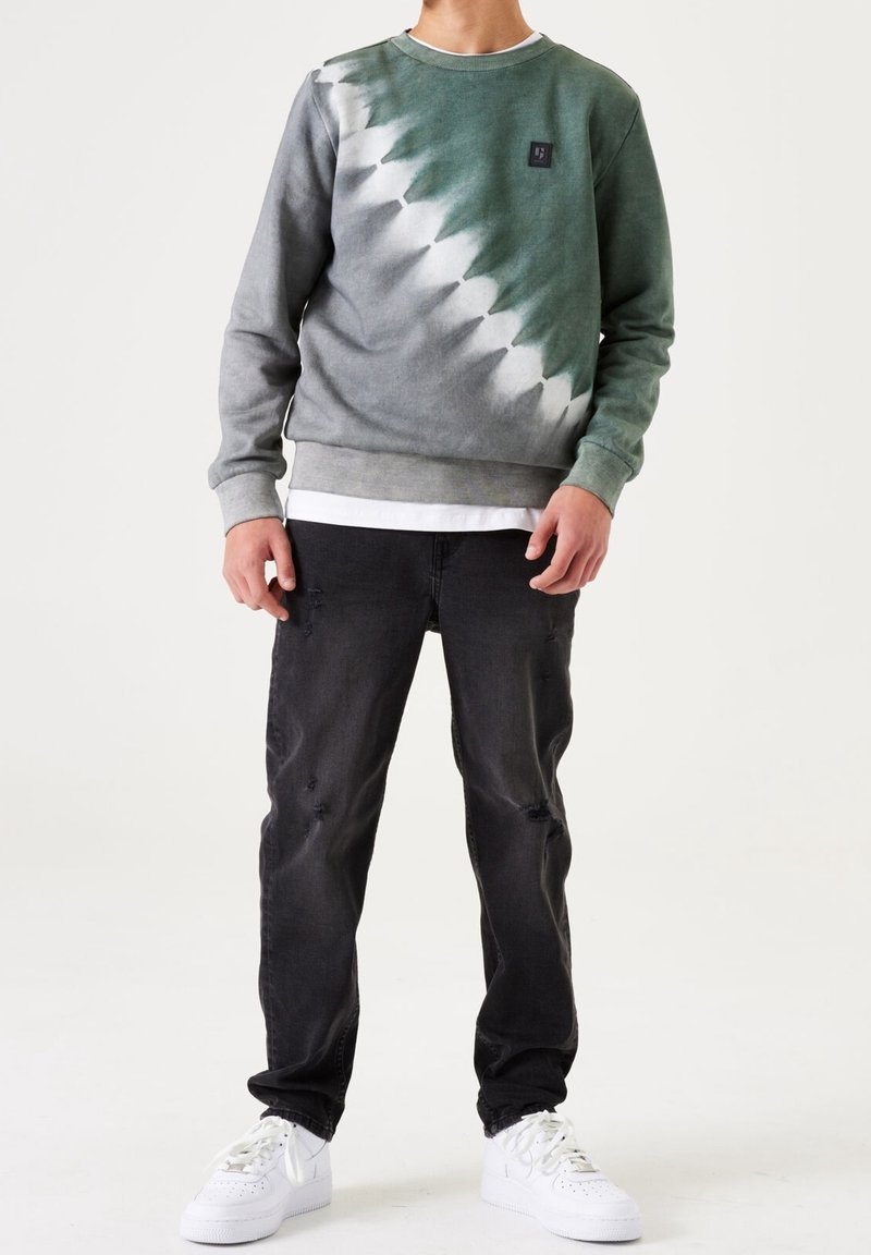 Felpa tie-dye verde e grigia con polsini e orlo a costine, caratterizzata da un piccolo logo nero. Abbinata a jeans neri strappati e sneakers bianche.