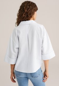 White shirt with light blue stripes, wide sleeves, a back pleat, and a classic collar. Paired with blue jeans.