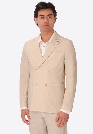 Uomo che indossa una giacca doppiopetto beige e pantaloni abbinati sopra una camicia bianca, in piedi contro uno sfondo semplice.