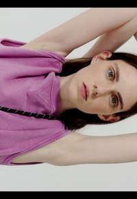 Persona giovane con pelle chiara e capelli castani che indossa un top viola senza maniche, alzando entrambe le braccia dietro la testa su uno sfondo neutro.