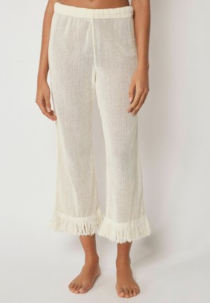 Frau trägt weiße, gehäkelte Hosen mit weitem Bein und Fransen am Saum, steht barfuß vor einem schlichten Hintergrund.