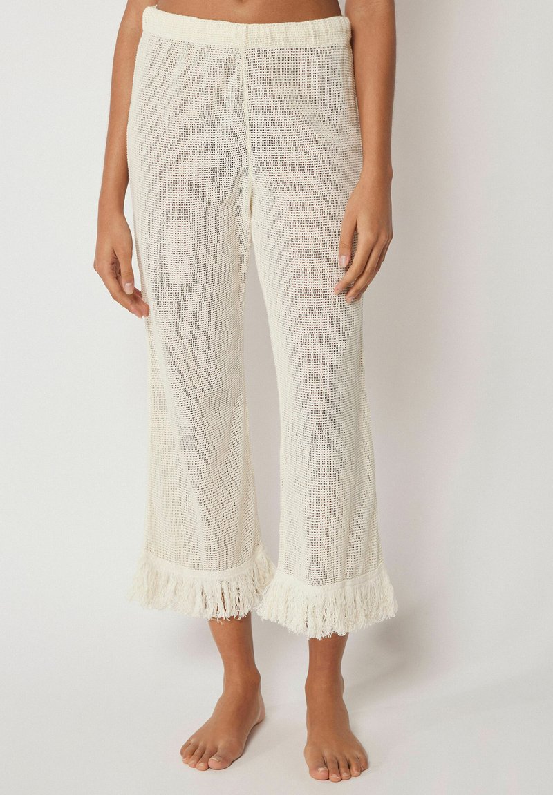 Mujer con pantalones anchos de crochet blancos con dobladillos con flecos, de pie descalza sobre un fondo liso.