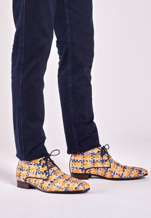 Geweven schoenen in oranje, blauwe en gele patronen; ronde neus, vetersluiting, platte hak, gecombineerd met donkere denimspijkerbroeken.