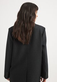 Blazer noir à rayures avec des épaules structurées et des manches longues, présentant une texture lisse et des accents de boutons sur les poignets.