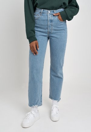 Osoba ubrana w jasnoniebieskie, wysokowaistowe spodnie jeansowe o prostych nogawkach z postrzępionymi krawędziami, ciemnozieloną bluzkę z długim rękawem, białe skarpetki i białe trampki.