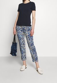 Marinblå t-shirt med kort ärm, blommiga byxor i blått och krämfärg, beige slip-on skor, bärande en marinblå väska med rynkad textur.