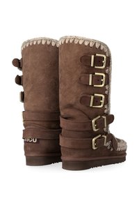 Stivali marroni in suede con rifiniture in pelliccia sintetica, caratterizzati da cinque fibbie dorate, dettagli intrecciati e una suola piatta. Texture morbida e design robusto.