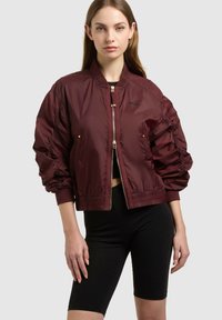 Femme portant une veste bomber bordeaux avec manches froncées et un short noir ajusté, debout devant un fond clair uni.