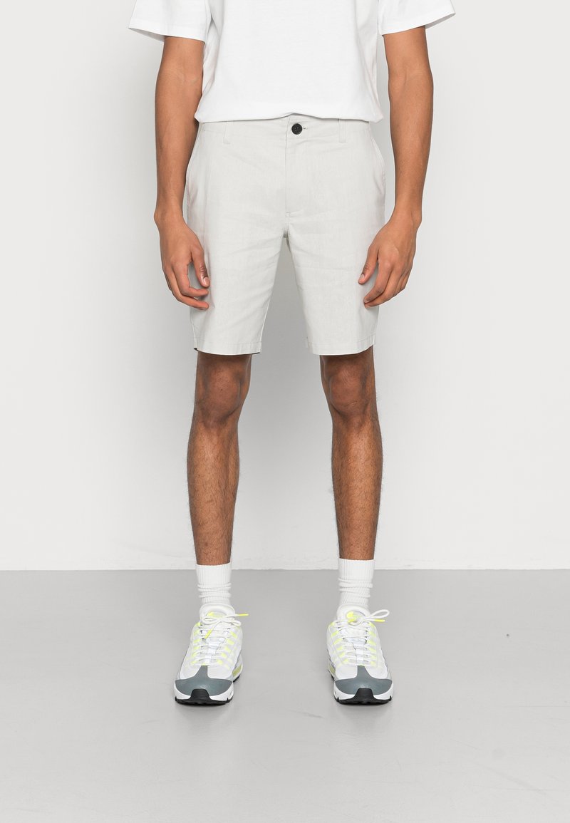Homme portant un short gris clair longueur genoux, des chaussettes blanches, des baskets blanches et grises avec des accents jaune néon et un t-shirt blanc.