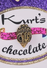 Metalowy pinezk z dekoracyjnym wzorem ptaka jest przymocowany do białej etykiety ozdobionej fioletowymi i różowymi akcentami brokatu oraz wyraźnym tekstem.