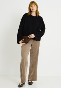 Suéter negro con textura, pantalones de pana marrón de pierna ancha y zapatos planos negros con tachuelas plateadas. Sosteniendo un bolso de mano de piel de serpiente marrón.