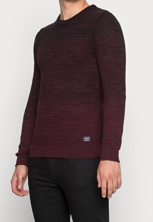 Mand iført en tætsiddende, mørk burgunderfarvet strikket sweater med ribstrikkede ærmekanter og kant forneden, kombineret med sorte bukser, stående mod en ensfarvet baggrund.