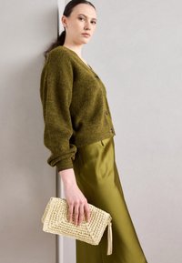 Cardigan en tricot vert olive avec fermeture à boutons, associé à une jupe olive en satin. Pochette tissée beige avec détail de pompon à la main.