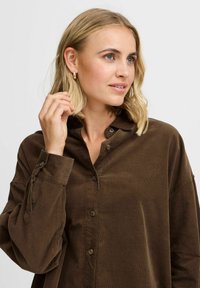 Camisa de pana marrón con cuello, cierre de botones en la parte delantera y mangas largas con puños atados. Textura suave con detalle de estrías verticales.